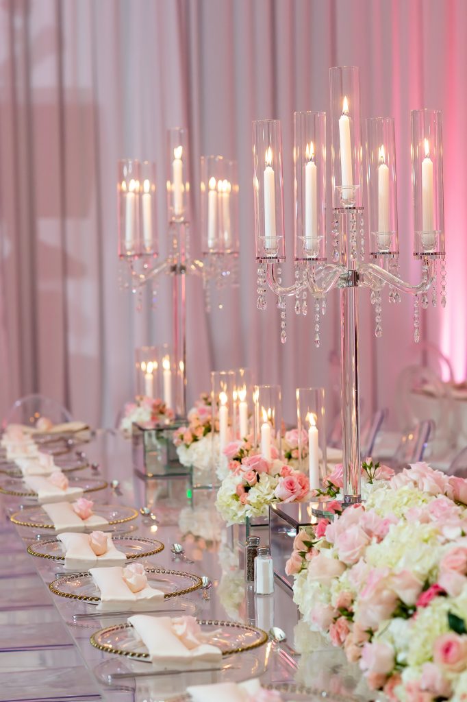 Image of a king's table with clear acrylic plates that have gold rims, and napkins on top. Low floral centerpieces in pink and white flow down the center of the table, and candles on mirrored boxes are interspersed throughout. Two tall candelabras are spaced down the center. 