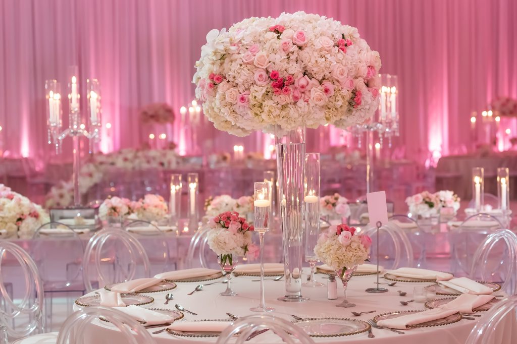 tall floral centerpiece on a tapered vase filled with a large centerpiece. There are small floral arrangements and other candles surrounding the main centerpiece. The table sits in front of a reception hall with other tables in the background with candelabras, and pink uplights on the walls. 